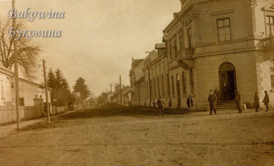 Wide unpaved street with a large corner building, men in coats on the steps and fir trees on the left