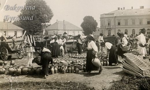 Market with rows of clay jugs on the ground, peasants in traditional dress, carts and a large building behind