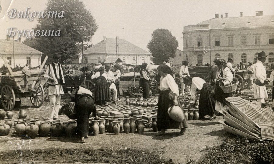 Market with rows of clay jugs on the ground, peasants in traditional dress, carts and a large building behind