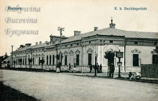 Long single-storey building with decorated windows, a noticeboard and people in peasant dress on the pavement