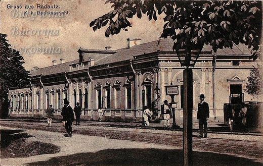 Long single-storey building seen past a street lamp and leafy branches, with men walking in front