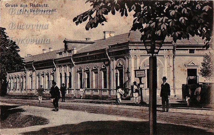 Long single-storey building seen past a street lamp and leafy branches, with men walking in front