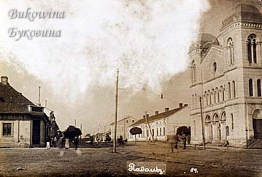 Street with low houses and people with umbrellas, and the tall towered front of a synagogue on the right