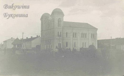 Large twin-towered temple with domed towers and arched windows beside low houses, an open square in front