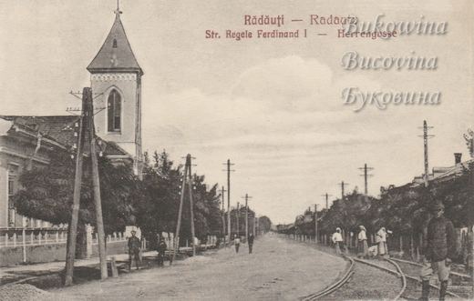 Long tree-lined street with rail tracks and telegraph poles, a pointed church tower at left and people walking