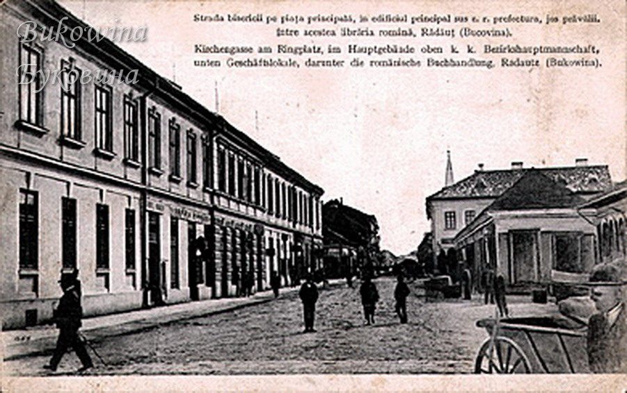 Cobbled street with a long two-storey building of shops at left, people walking and a cart in the foreground