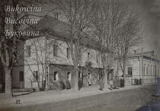 Two-storey house behind bare trees on a sunlit street, soldiers in greatcoats standing on the pavement