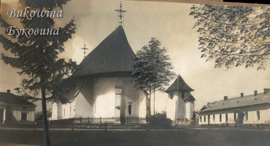 Church with a tall conical roof and Orthodox cross, a separate bell tower and long low building at right