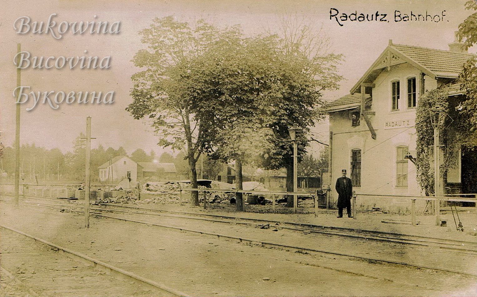 Station building signed Radautz beside the tracks, a uniformed man on the platform and trees with rubble behind