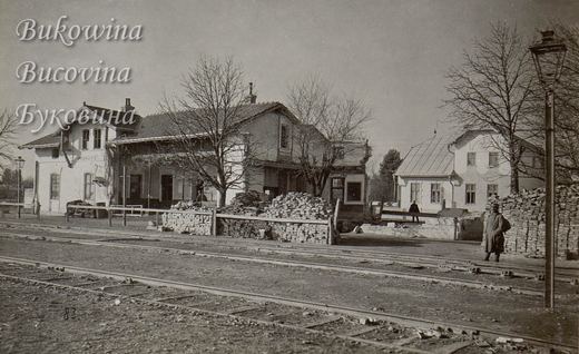 Station buildings beside the tracks with stacks of bricks and bare trees, a soldier standing by a lamp post