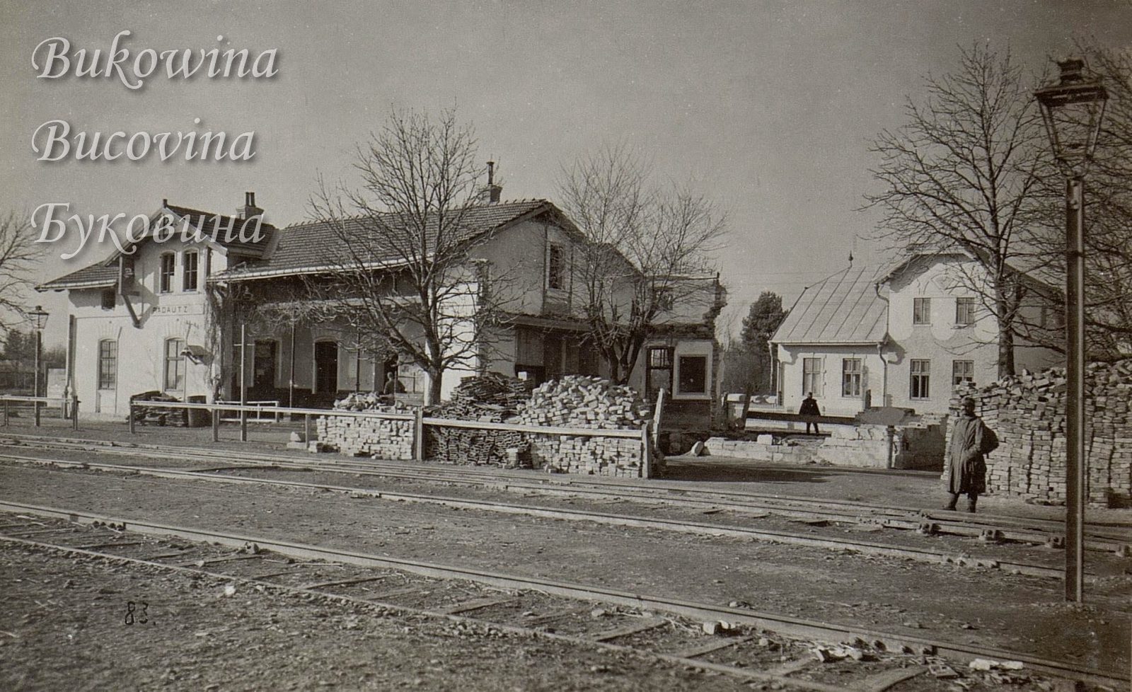 Station buildings beside the tracks with stacks of bricks and bare trees, a soldier standing by a lamp post