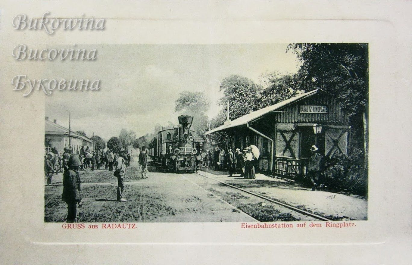 Small steam train at a wooden station signed Radautz-Ringplatz, crowds and a woman with a parasol nearby