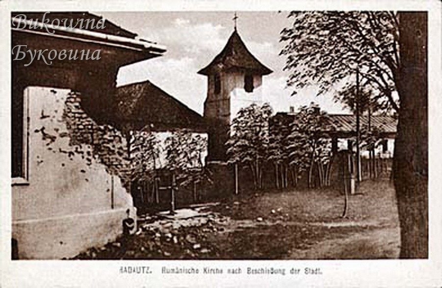 Damaged house wall with exposed brick at left, a church bell tower and trees in a churchyard behind