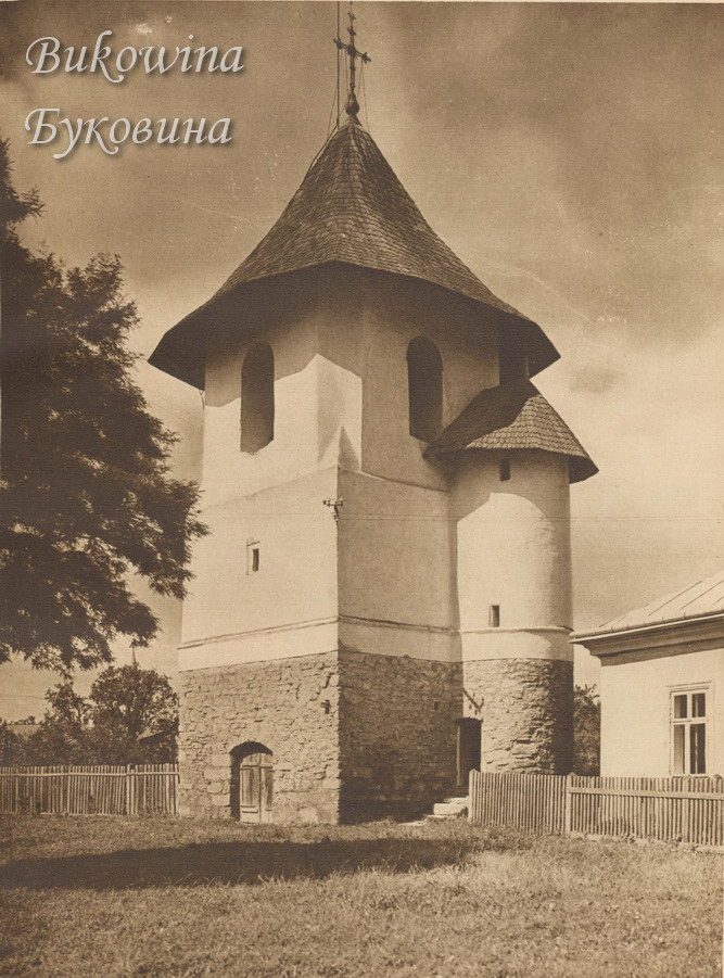 Stone and white-plastered bell tower with a shingled roof, a round stair turret and a cross, behind a fence