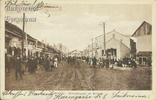 Muddy street crowded with people by shops and houses, telegraph poles, handwriting along top and bottom