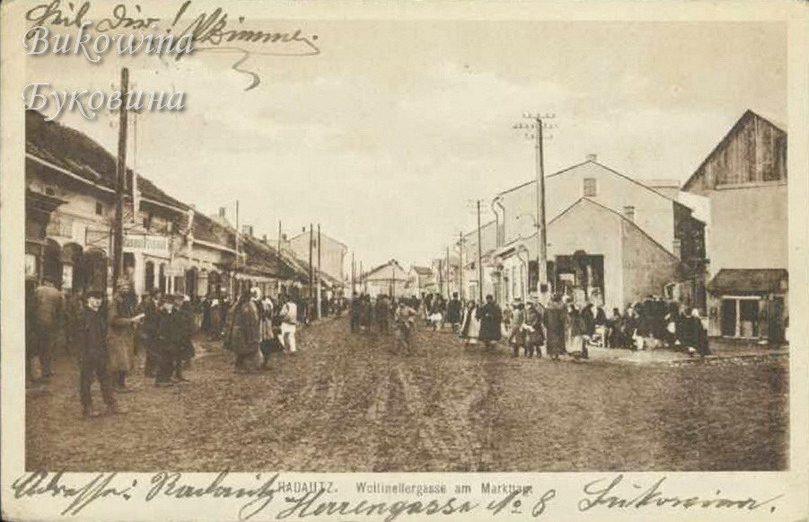 Muddy street crowded with people by shops and houses, telegraph poles, handwriting along top and bottom