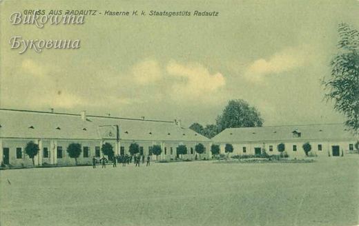 Long low barrack buildings with small trees around a wide empty yard, a few men standing near the buildings