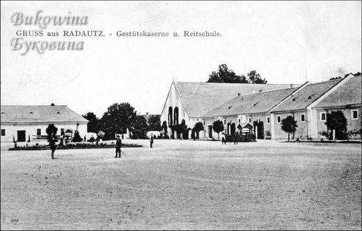 Wide open yard before a tall-gabled riding hall and long barrack buildings, a few men standing in the yard