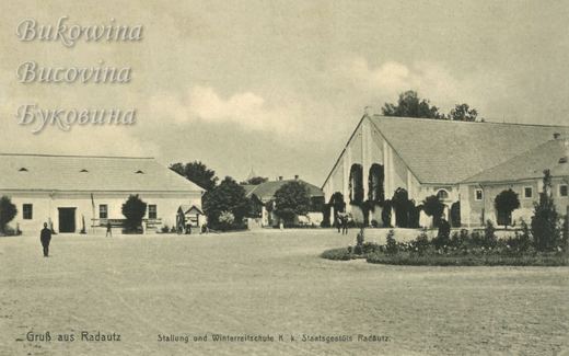 Big yard with a flower bed before a gabled riding hall with ivy-covered arched windows and long stables