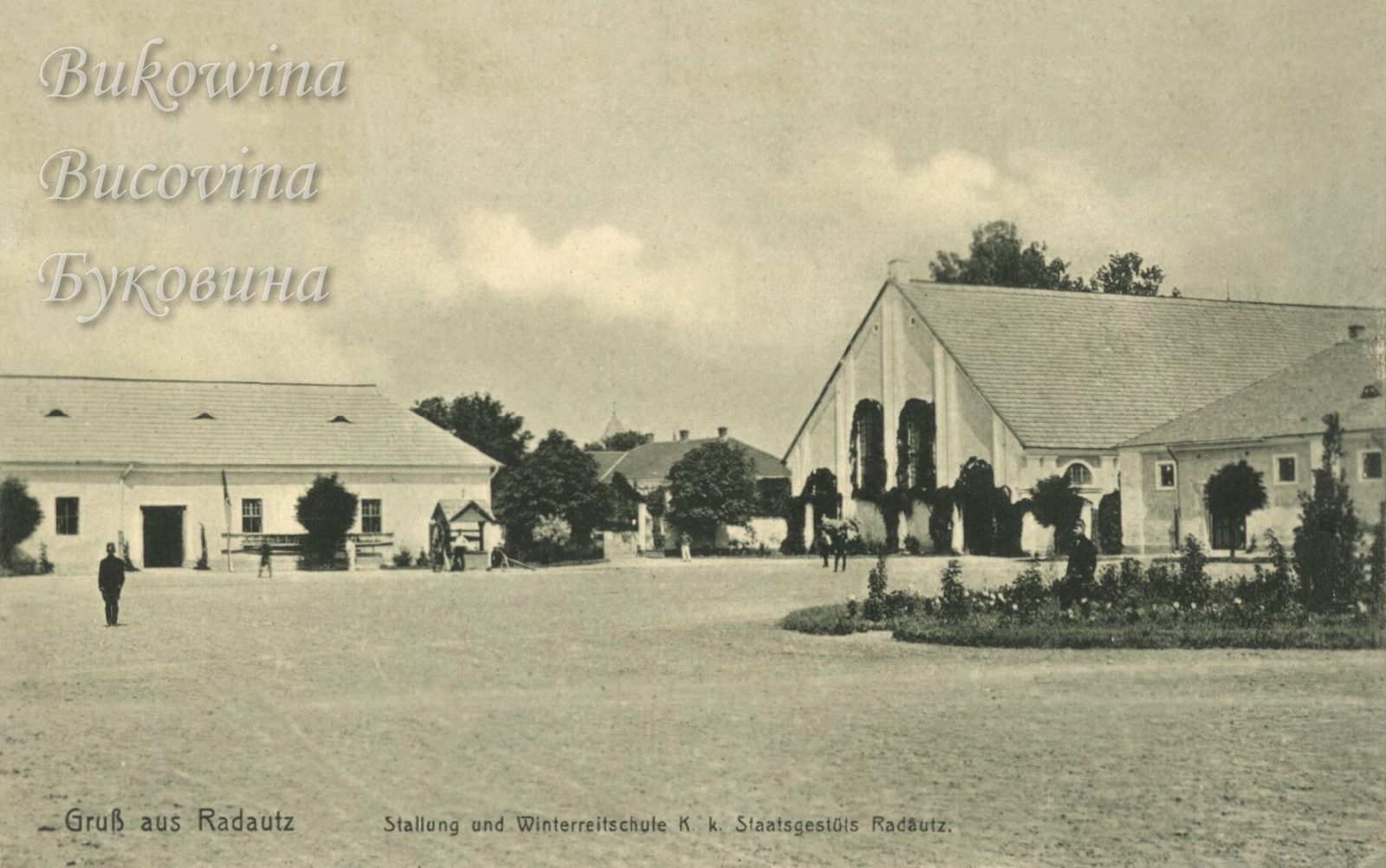 Big yard with a flower bed before a gabled riding hall with ivy-covered arched windows and long stables