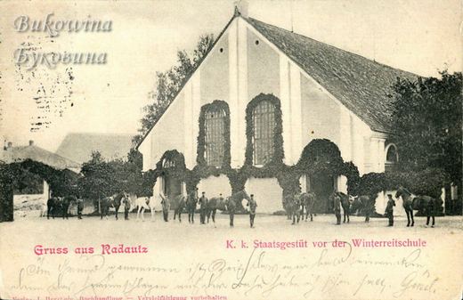 Men holding a line of horses in front of a gabled riding hall with ivy-framed windows, handwriting below