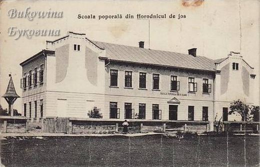 Two-storey school building with gabled end wings behind a wooden fence, one person standing in front