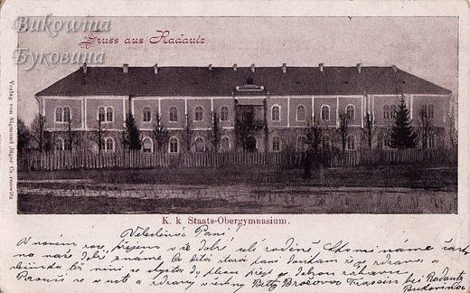 Long two-storey school building with arched windows behind a picket fence, with a handwritten message below