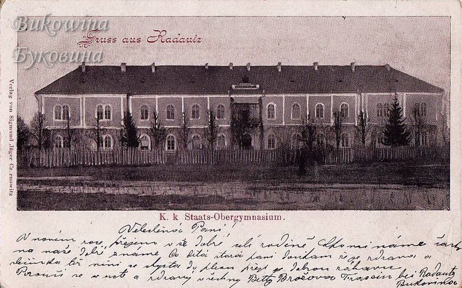 Long two-storey school building with arched windows behind a picket fence, with a handwritten message below