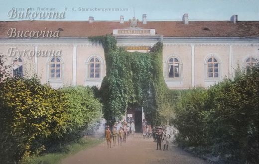 Ivy-covered entrance of a school marked Franz Josef I, with boys and men standing on a path between bushes
