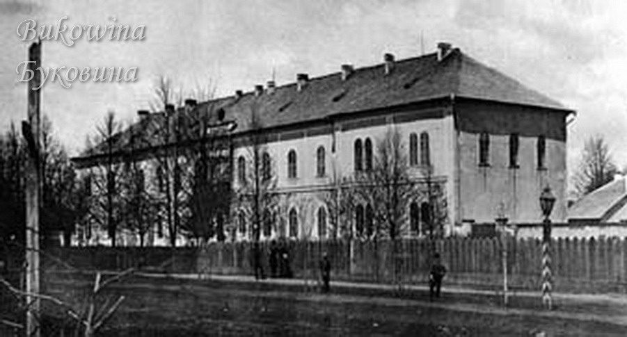Large two-storey building behind a wooden fence, with bare trees, a street lamp and a few people on the road