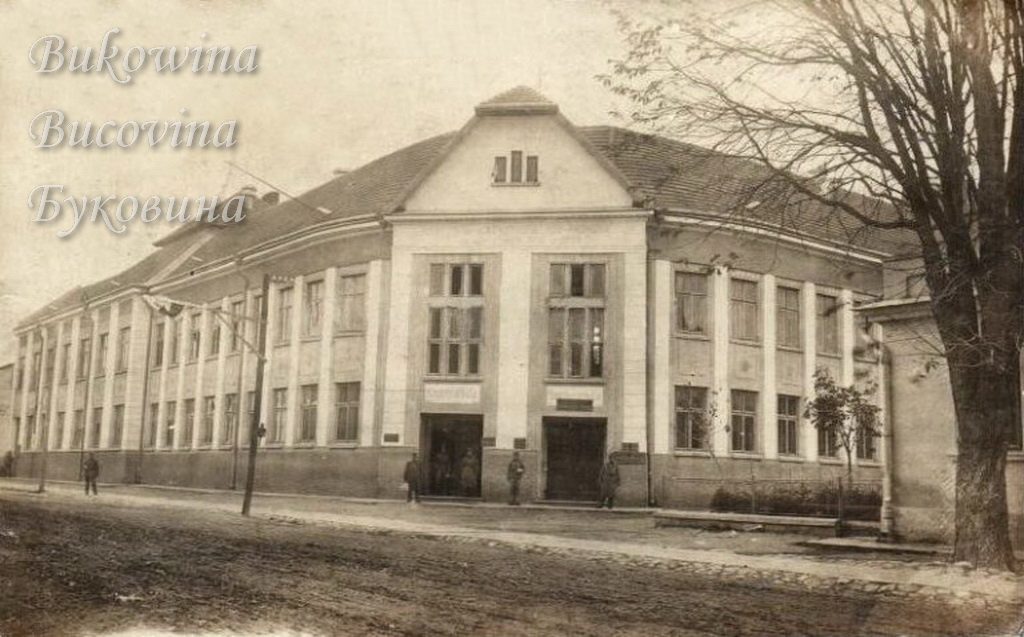 Large two-storey building with a central gable and two entrances on a curving street, a bare tree on the right