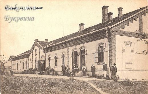 Long single-storey brick-trimmed factory building with barrels by the door and a row of men in hats in front