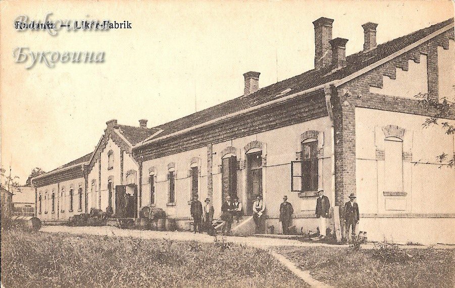 Long single-storey brick-trimmed factory building with barrels by the door and a row of men in hats in front
