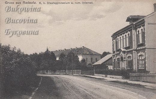 Unpaved street with an ornate two-storey building on the right and a large school building behind trees