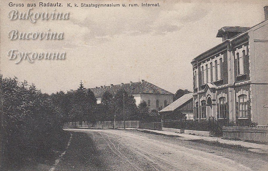 Unpaved street with an ornate two-storey building on the right and a large school building behind trees