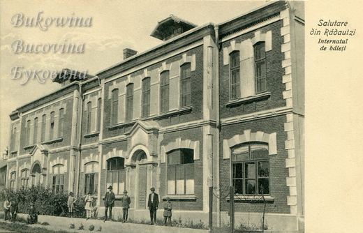 Two-storey brick building with arched windows and stone trim, men and boys standing along the pavement in front