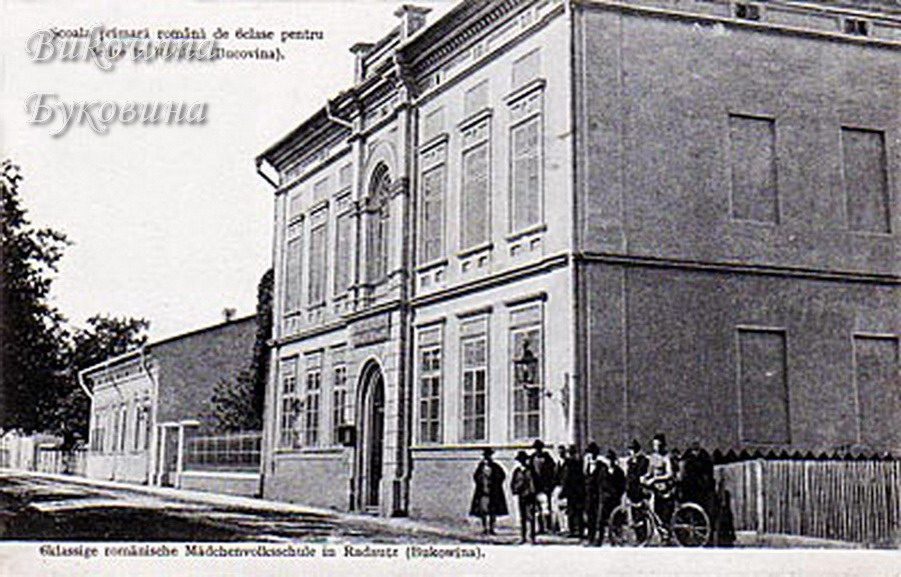 Two-storey corner school building on a street, a group of people and a man with a bicycle in front of the fence
