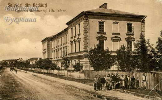 Long three-storey barracks beside a dirt road, young trees and a group of people standing by a wooden fence