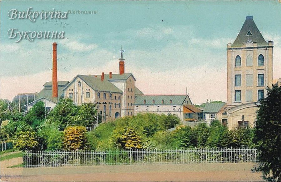 Coloured view of brewery buildings with tall chimneys and a square tower, behind bushes and an iron fence