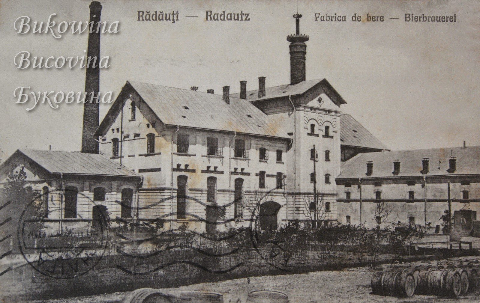 Large brewery complex with a tall chimney and a tower, rows of wooden barrels in the yard, postmarks across the front