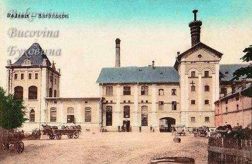 Coloured view of a brewery yard with an arched gateway, a tall tower and horse carts loaded with barrels