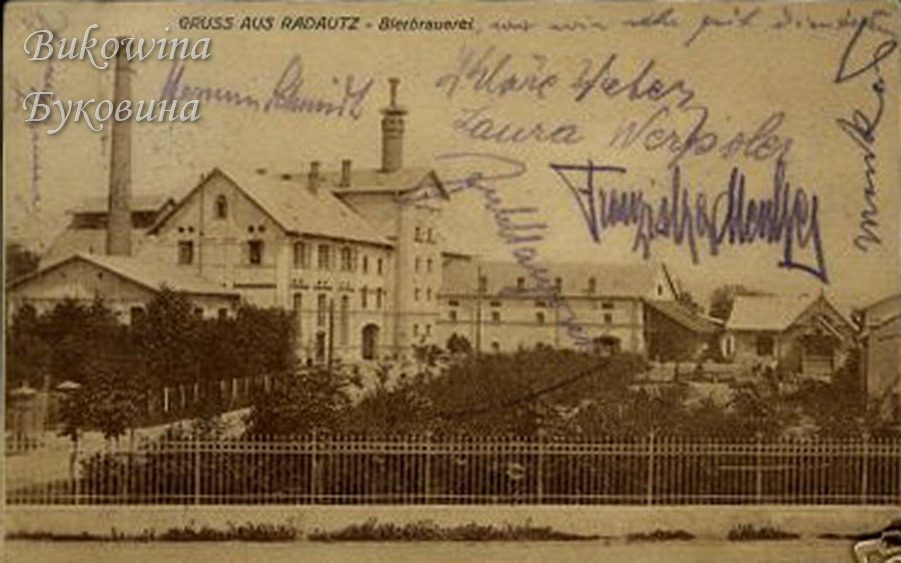 Brewery buildings with a tall chimney behind trees and an iron fence, several handwritten signatures across the sky