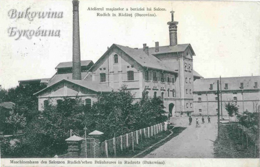 Brewery engine house with a tall chimney and tower, a lane with people walking and trees behind a fence