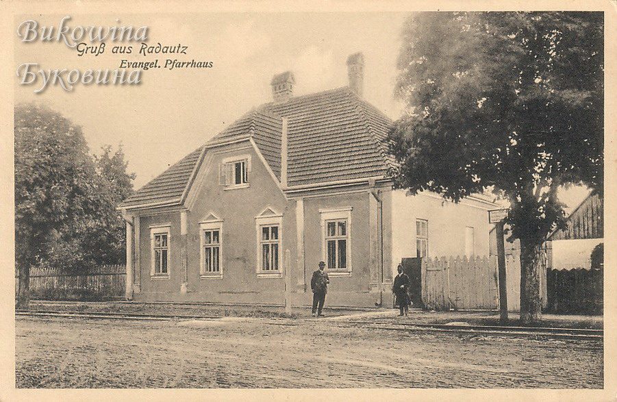 Single-storey house with a tiled hipped roof and gabled front, two men standing on the street beside a large tree