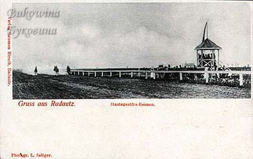 Racecourse with a white rail fence, spectators and a raised pavilion with a pennant, riders in the distance
