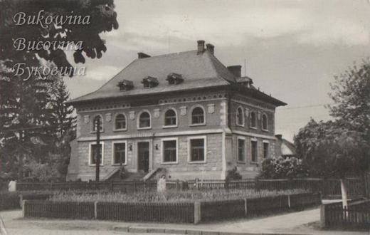 Two-storey villa with arched upper windows and dormers in a hipped roof, behind a fenced front garden