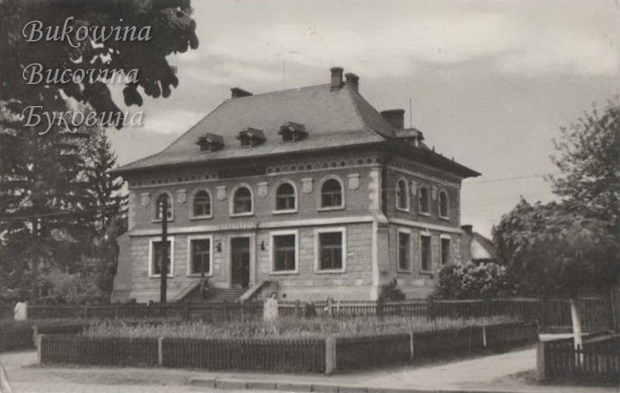 Two-storey villa with arched upper windows and dormers in a hipped roof, behind a fenced front garden