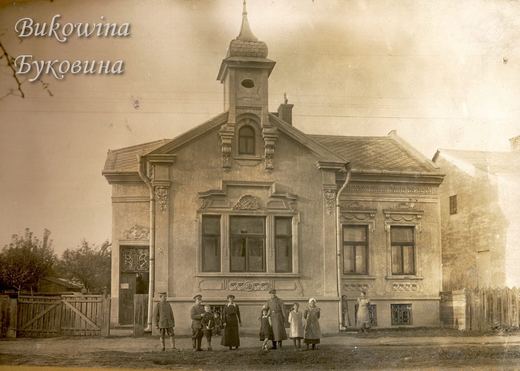 Single-storey house with stucco ornament and a small domed turret, a family and soldiers standing in front