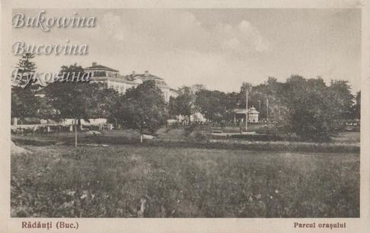 Grassy park with trees, a small pavilion and a large two-storey building in the background
