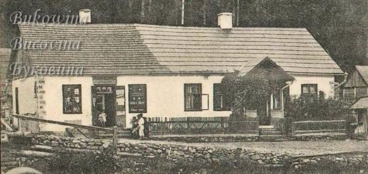 Long whitewashed single-storey house with a shingle roof, open shop door, picket fence and stone wall, forest behind
